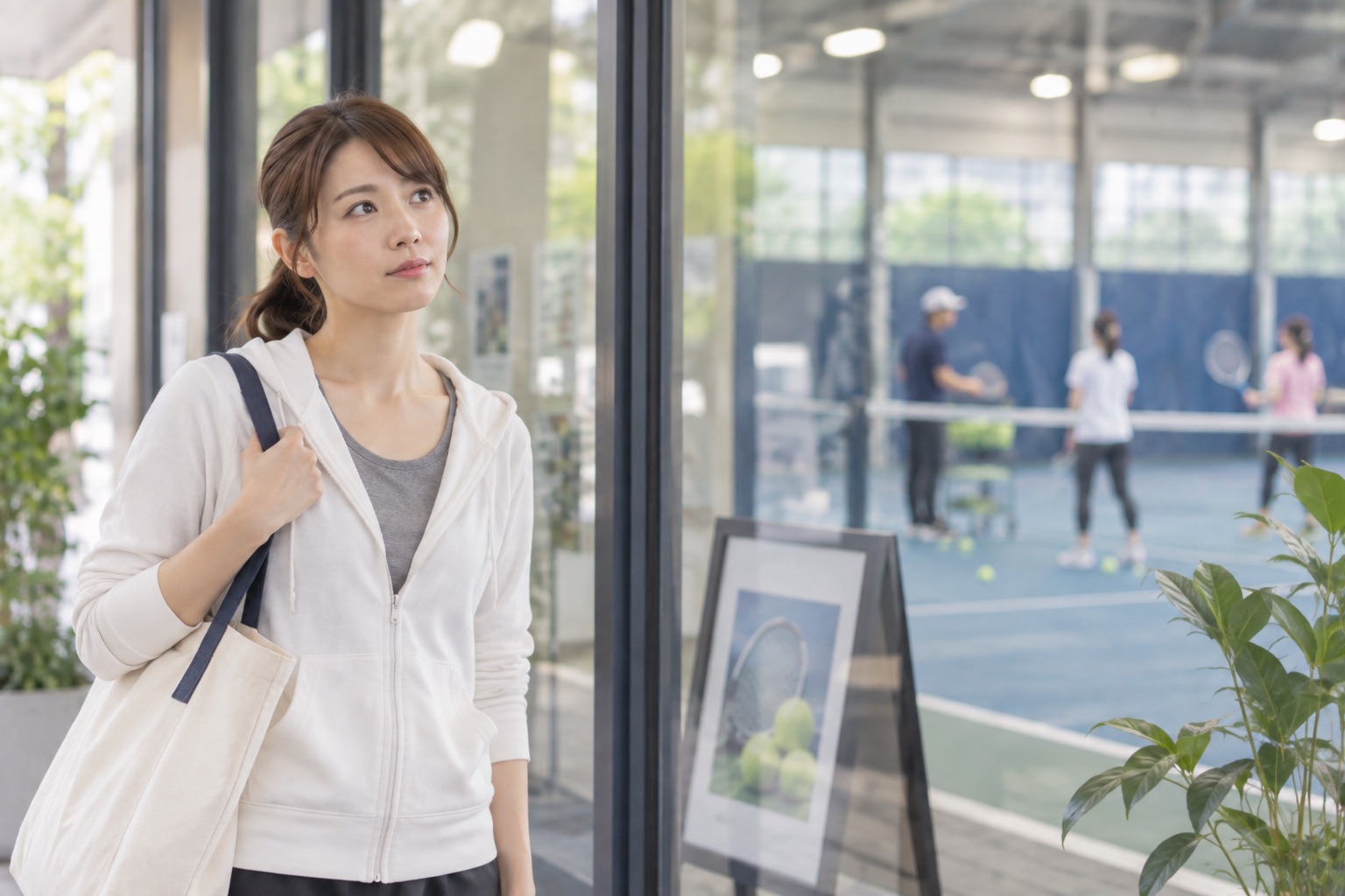 バッグを持った女性が、テニススクールの入口から屋内コートでレッスンを受ける人たちを外側から見ている